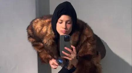 Winter coats for women featuring a woman in a brown faux fur coat, black hoodie, and holding a smartphone.